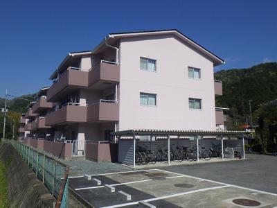 手前に駐車場と駐輪場があるピンク色で3階建ての建物の写真
