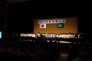 平成31年丹波市成人式と書かれた垂れ幕のあるホールの舞台で合唱する人たちを正面から撮影した写真