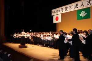 平成31年丹波市成人式と書かれた垂れ幕のあるホールの舞台で合唱する人たちを舞台右側から撮影した写真