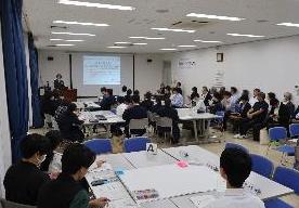 高校生、大学生、議員が班ごとに円を作り、発表者の発表を聞いている写真