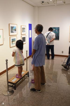 小さい台に乗った子供と後ろで見守っている親が一緒に展示物を見ている写真