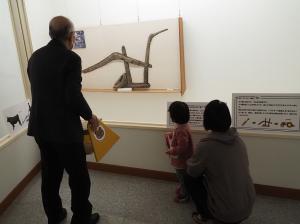 展示物の前に立っているスーツ姿の男性とその横で展示物を眺めている親子の写真