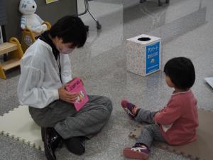 小さなマットを床に敷いて向かい合って座る、絵本を手にした女性と小さな女の子の写真