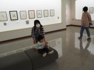 絵画の展示される美術館内のソファに座る子どもと、付きそう親の写真