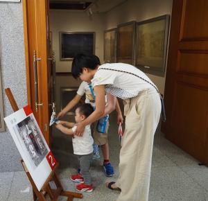 親と年上の子の助けを借りながら、美術館内の扉の取っ手にくくりつけられた物に手を伸ばす幼児の写真