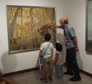 黄色い葉が繁る森が描かれた、大きな絵の一部分を指差す男性と、見上げる二人の子どもの写真