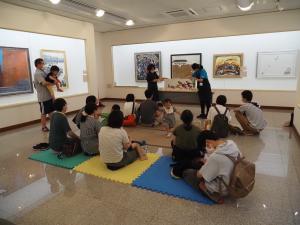 絵画の展示室で絵本の読み聞かせを聞くたくさんの親子の写真