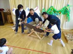 子どもが大人と一緒に、のこぎりで木を切る体験をしています。