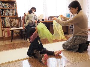 小さな女の子と美術館職員が、スカーフを使って歌と手遊びを楽しんでいます。その様子をお母さんが笑顔で見ています。