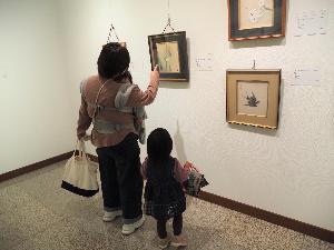 小さな女の子がお母さんと赤ちゃんと一緒に絵を見ています。お母さんが絵を指さして蝶々がいるよと話しかけています。