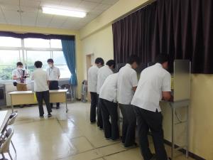 記載台を使って記入している生徒と、奥の投票箱に投票する男子生徒たちの写真